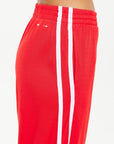 A person is wearing The Upside's Juliet Pant, which is red with white side stripes. These low-rise pants are made from a soft double-faced jersey and include an elastic waistband, along with a small white logo near the hip. The background is plain white.