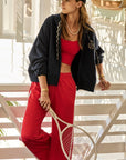 A woman dressed in a casual athletic ensemble leans against a railing, showcasing her icy red crop top paired with The Upside's matching low-rise Juliet Pant. She holds a tennis racket and accessorizes with layered necklaces and bracelets as sunlight streams in behind her.