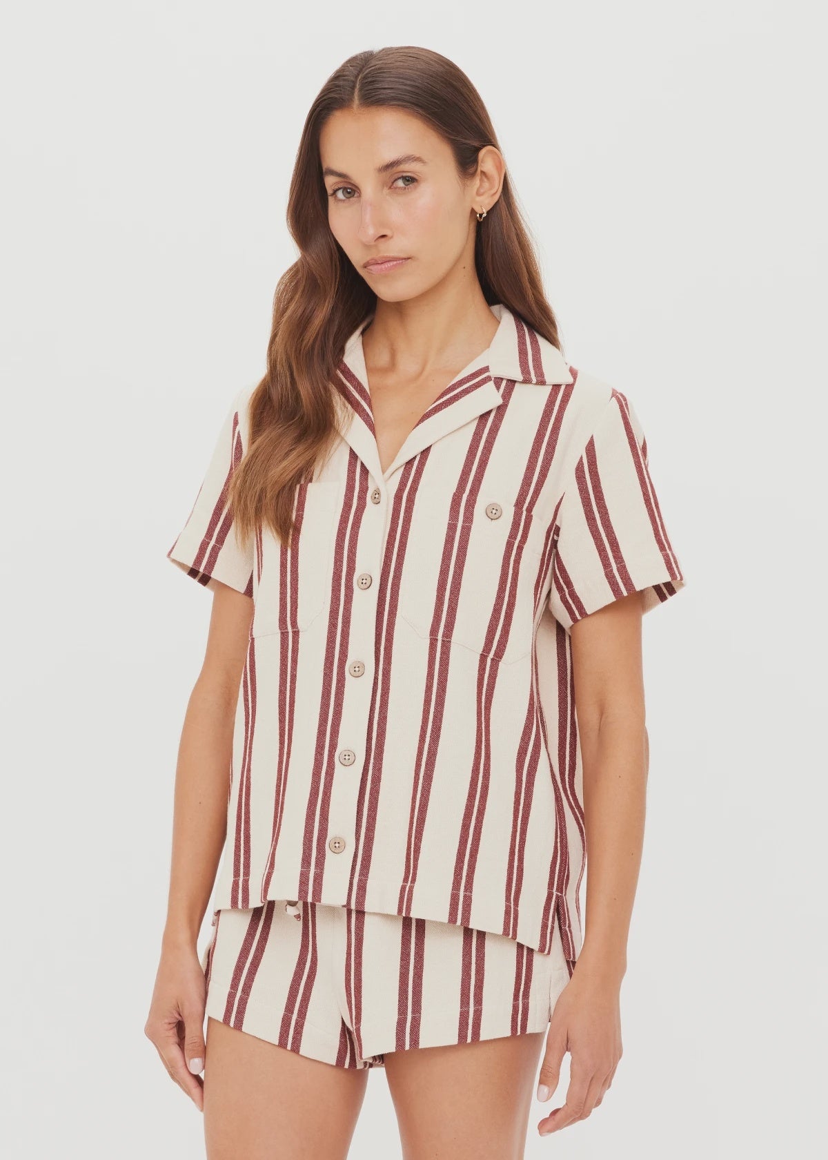 A woman with long brown hair and a neutral expression wears The Upside’s BAJA KATIA COLLARED SHIRT, a short-sleeve striped button up shirt and shorts set with vertical maroon stripes on cream, standing against a plain white background.