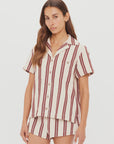 A woman with long brown hair and a neutral expression wears The Upside’s BAJA KATIA COLLARED SHIRT, a short-sleeve striped button up shirt and shorts set with vertical maroon stripes on cream, standing against a plain white background.