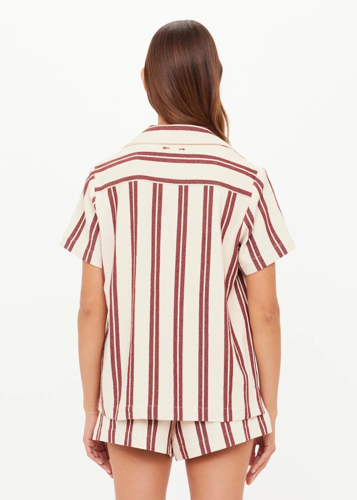A woman with long brown hair faces away, wearing The Upside's BAJA KATIA COLLARED SHIRT—an organic cotton, short-sleeve, cream top with vertical maroon stripes. She is against a plain white background.