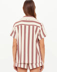A woman with long brown hair faces away, wearing The Upside's BAJA KATIA COLLARED SHIRT—an organic cotton, short-sleeve, cream top with vertical maroon stripes. She is against a plain white background.