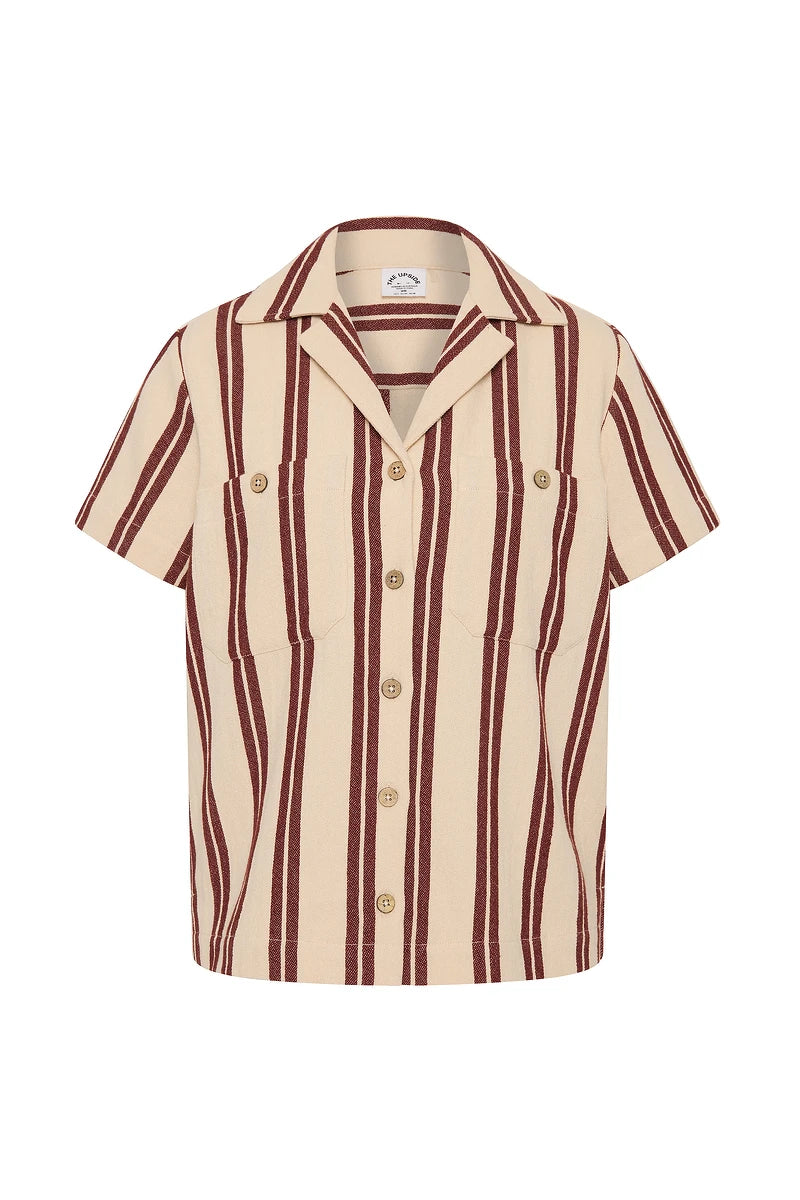 The Upside’s BAJA KATIA COLLARED SHIRT is a short-sleeved button-up with wide vertical burgundy stripes on beige, two chest pockets, and a classic collar—ideal for effortless coastal style.