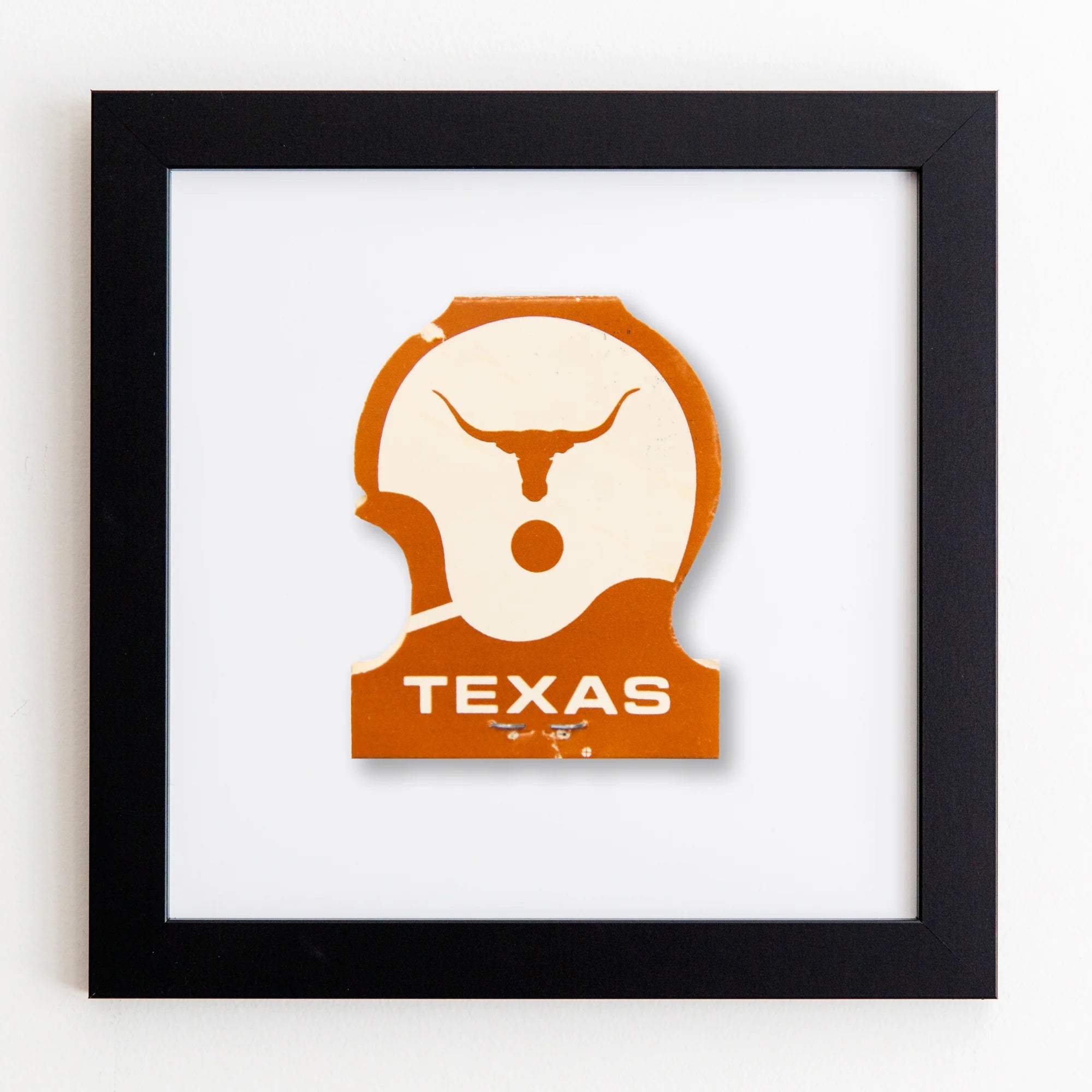 A framed artwork featuring a stylized silhouette of a longhorn&#39;s head in white acrylic on an orange background with &quot;TEXAS&quot; written below, all enclosed in a Match South Art Square Black Frame.