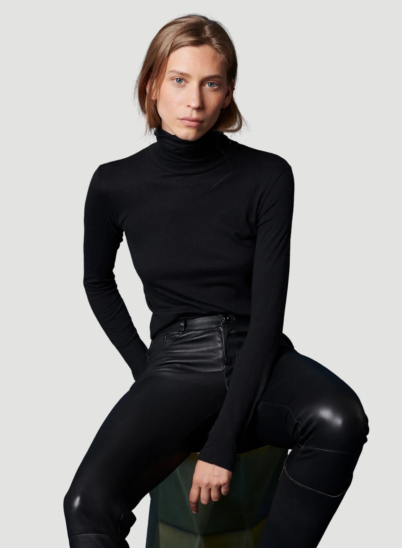 A person with light brown hair sits on a stool, wearing the Majestic Filatures Soft Touch L/S Turtleneck in black and black leather pants, confidently looking at the camera against a plain light gray background.