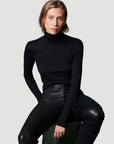 A person with light brown hair sits on a stool, wearing the Majestic Filatures Soft Touch L/S Turtleneck in black and black leather pants, confidently looking at the camera against a plain light gray background.