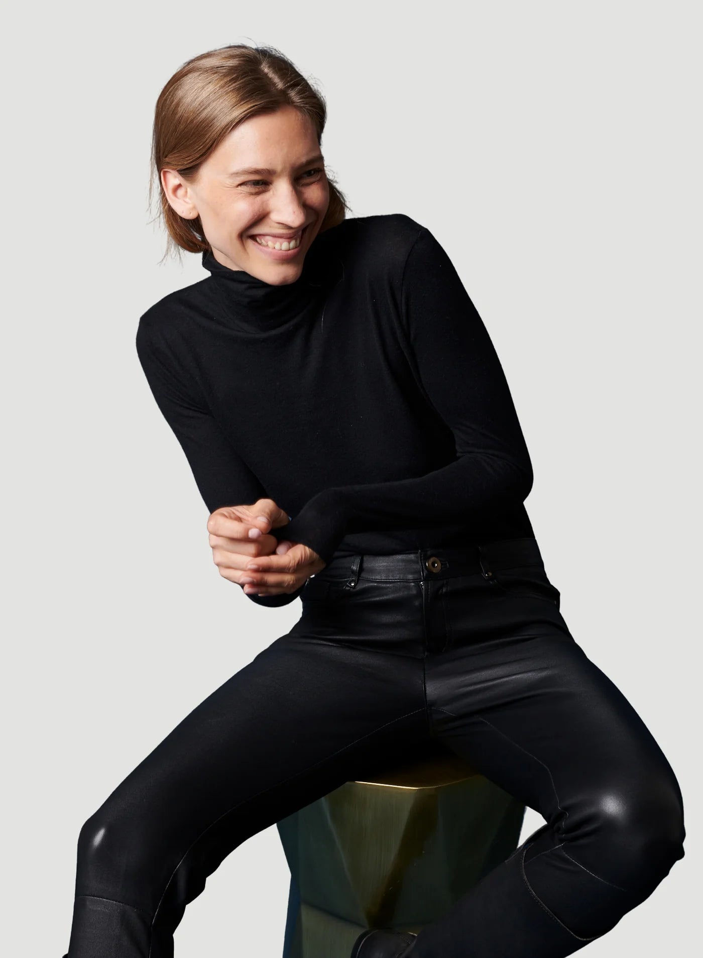 A person with light brown hair, wearing a Majestic Filatures Soft Touch L/S Turtleneck and black leather pants, sits on a stool and smiles, looking to the side against a plain light gray background.