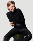 A person with light brown hair, wearing a Majestic Filatures Soft Touch L/S Turtleneck and black leather pants, sits on a stool and smiles, looking to the side against a plain light gray background.
