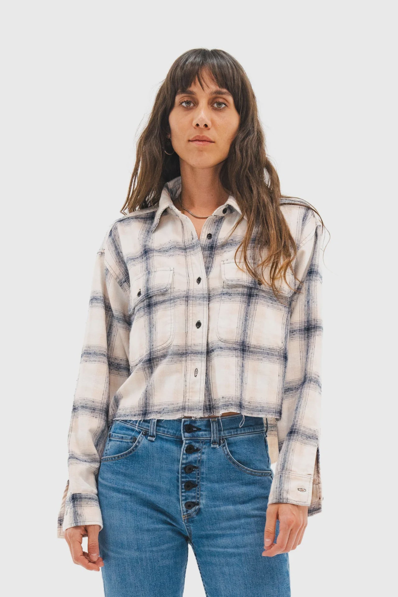 A woman with long brown hair wears the ASKK Crosby Flannel, a white and blue plaid button-up shirt, paired with high-waisted blue jeans, standing facing forward against a plain light background.