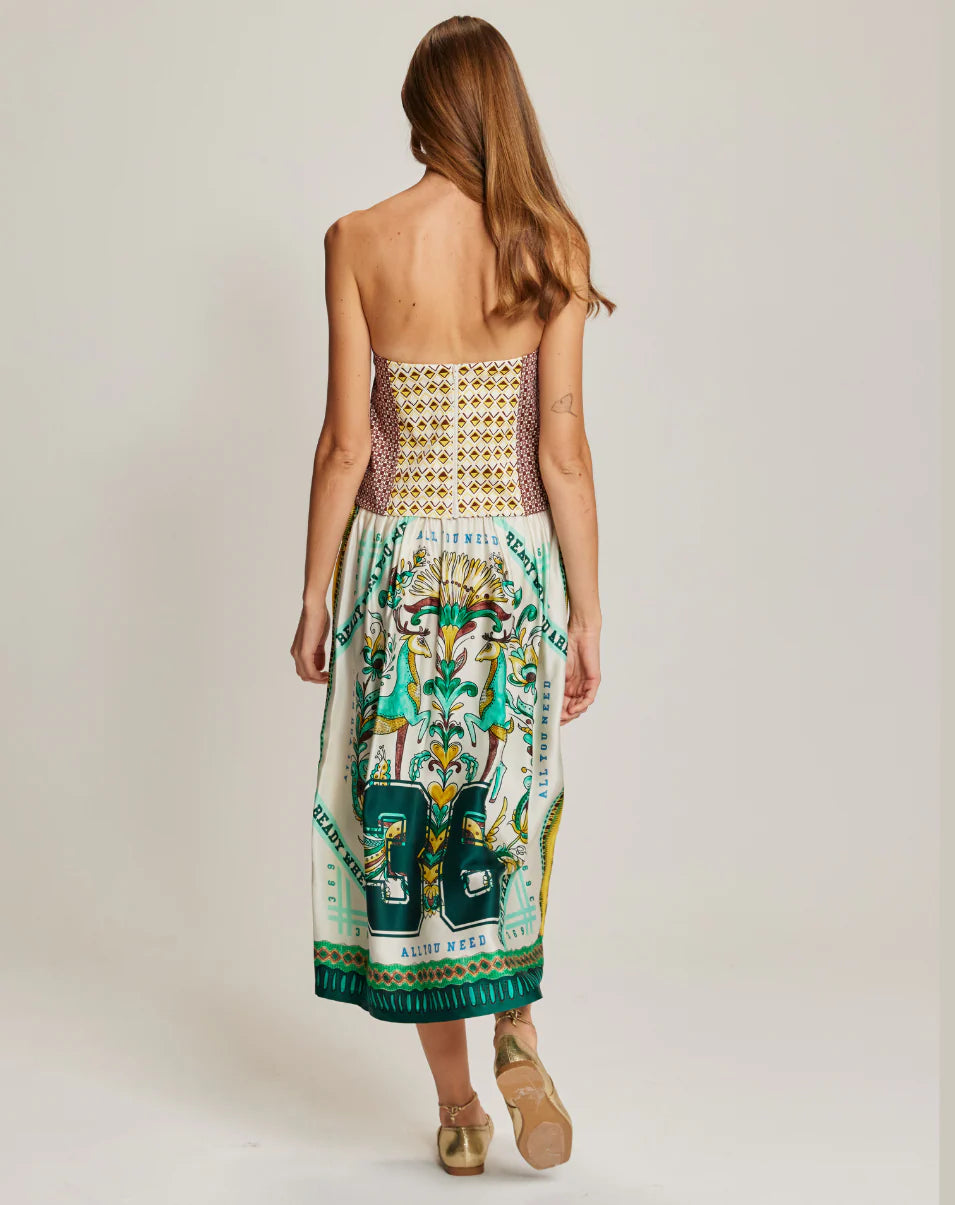 A woman with long brown hair faces away, wearing the Me369 Vanessa Midi Skirt—a colorful illustrated viscose midi skirt with an elastic waist and a fitted, patterned bodice. She pairs it with gold shoes against a plain light background.
