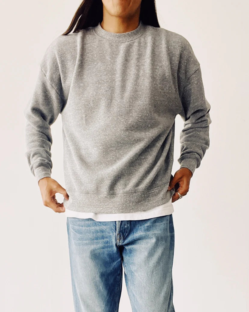A person with long hair wears The Boxy Sweatshirt by Hey Gang, styled in an oversized vintage look, layered over a white shirt and blue jeans, standing in a neutral pose against a light background.