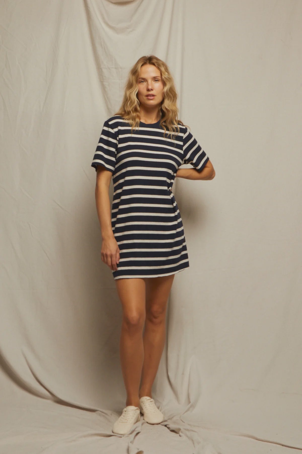 A blonde woman poses confidently with one hand on her hip, wearing the Rhett SS Tee Dress by Perfectwhitetee— a loose recycled cotton T-shirt dress— paired with white sneakers against a draped backdrop.