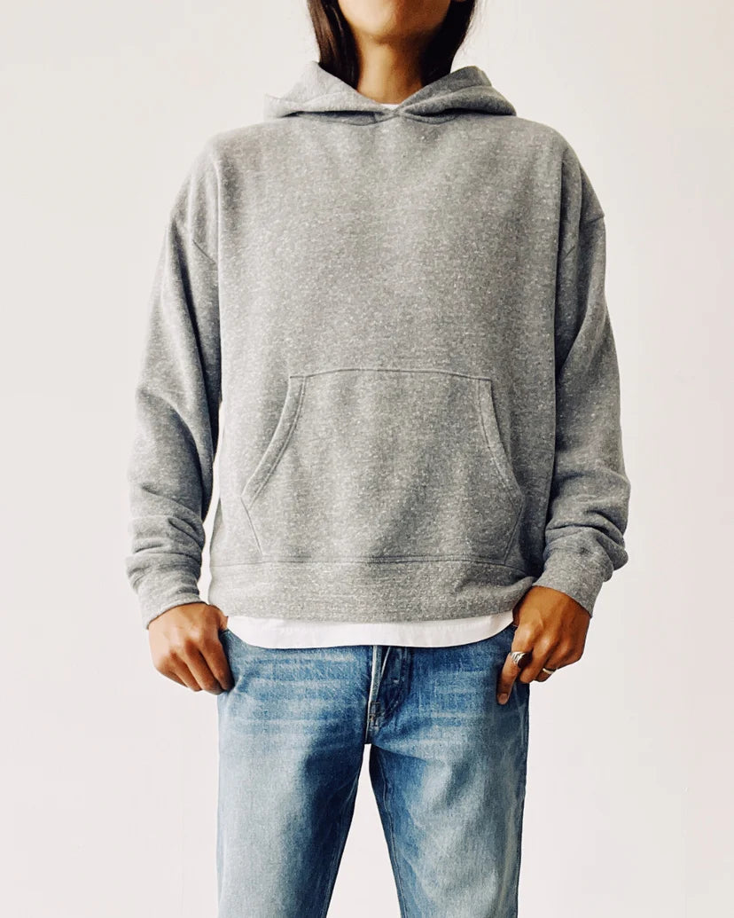 A person wearing The Hoodie from Hey Gang, designed in a gray shade, paired with blue jeans is standing against a plain, light background. The hoodie offers a boxy fit with unisex sizing and includes a front pocket where their hands rest casually.