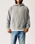 A person wearing The Hoodie from Hey Gang, designed in a gray shade, paired with blue jeans is standing against a plain, light background. The hoodie offers a boxy fit with unisex sizing and includes a front pocket where their hands rest casually.