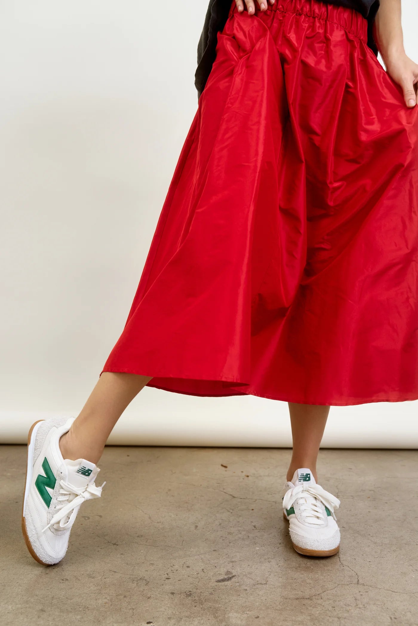 A person in an Aquarius Cocktail&#39;s red LE SKIRT Silk Taffeta midi skirt and white sneakers with green accents exudes style on a concrete floor, blending a modern take on 40&#39;s elegance with timeless charm in today&#39;s fashion scene.