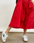 A person in an Aquarius Cocktail's red LE SKIRT Silk Taffeta midi skirt and white sneakers with green accents exudes style on a concrete floor, blending a modern take on 40's elegance with timeless charm in today's fashion scene.