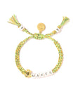 The Venessa Arizaga Bracelet 6" is a green and yellow braided charm bracelet with white beads spelling "MAKER," a gold charm, tassels, and an adjustable knot closure—ideal for fans of quirky jewelry.