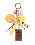 The Venessa Arizaga Bag Key Leash features playful food charms—a chocolate bar, bundt cake, yellow fry—plus a yellow sponge, bottle, purple beads, and "VIBES" letter beads on a silver keyring. Fun as a whimsical bag charm!.