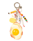 The Venessa Arizaga Bag Key Leash is a playful keychain bag charm with a large fried egg, cigarette, mushroom, yellow sponge, and colorful "NOT FRIED" beads—all attached to a sturdy metal key ring.
