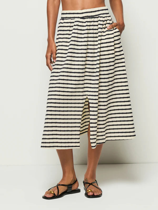 A person wears Nation LTD's Vida Skirt, a high-waisted black and white striped midi crafted from soft Peruvian cotton, featuring an elastic waistband and center front slit. They effortlessly pair it with black strappy sandals against a plain white backdrop.