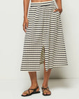 A person wears Nation LTD's Vida Skirt, a high-waisted black and white striped midi crafted from soft Peruvian cotton, featuring an elastic waistband and center front slit. They effortlessly pair it with black strappy sandals against a plain white backdrop.