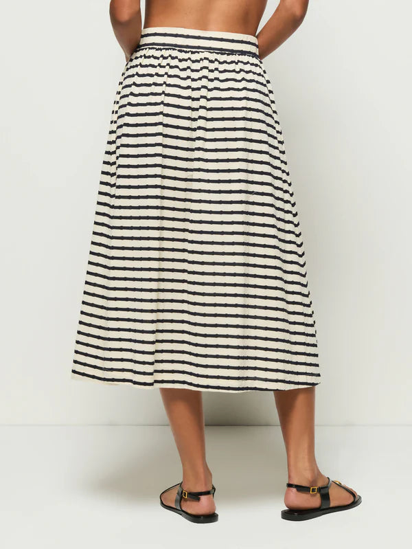 In the photo, a person stands barefoot against a white backdrop, wearing the Vida Skirt by Nation LTD. This high-waisted, calf-length skirt is crafted from soft Peruvian cotton and features beige and black stripes, an elastic waistband, a relaxed fit, and a subtle center front slit.