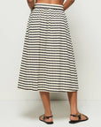 In the photo, a person stands barefoot against a white backdrop, wearing the Vida Skirt by Nation LTD. This high-waisted, calf-length skirt is crafted from soft Peruvian cotton and features beige and black stripes, an elastic waistband, a relaxed fit, and a subtle center front slit.