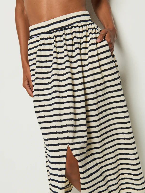 A person wears the Vida Skirt by Nation LTD, crafted from Peruvian cotton, showcasing black and white horizontal stripes. The skirt features a center front slit, an elastic waistband, and pockets; they stand against a plain background with one hand casually in a pocket.