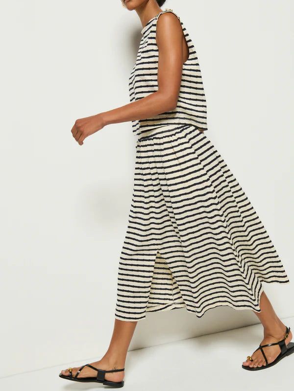 A person wearing the Nation LTD Vida Skirt, a black and white sleeveless striped dress with an elastic waistband, walks to the left in black sandals against a plain white wall.