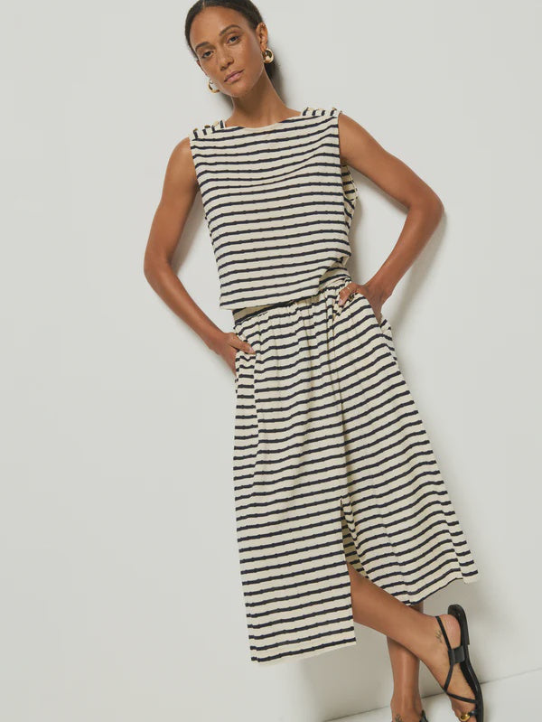 A person stands against a white background, wearing a sleeveless black and white striped top and the chic Vida Skirt by Nation LTD, crafted from soft Peruvian cotton. With one hand in their pocket, they accessorize with black sandals and gold hoop earrings.