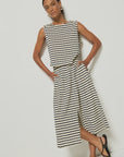 A person stands against a white background, wearing a sleeveless black and white striped top and the chic Vida Skirt by Nation LTD, crafted from soft Peruvian cotton. With one hand in their pocket, they accessorize with black sandals and gold hoop earrings.