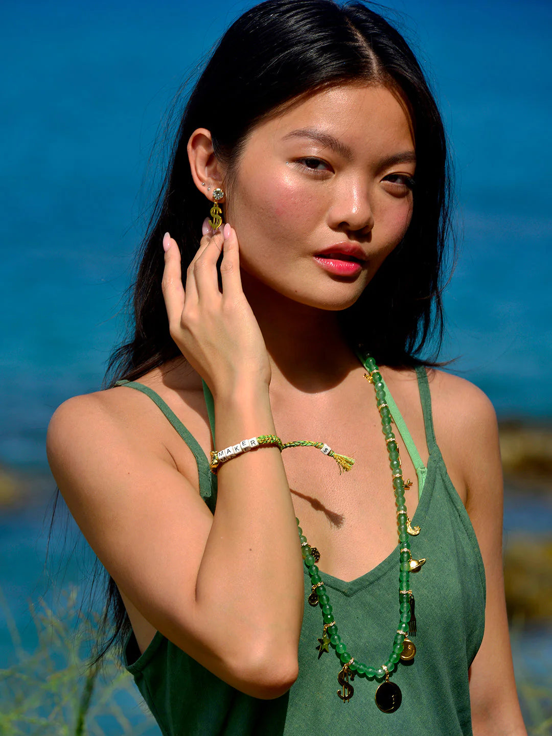 A woman with long dark hair stands by the water, wearing a green dress and accessorized with a Venessa Arizaga Bracelet 6", along with green and gold jewelry. She touches her earring and gazes at the camera.