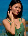 A woman with long dark hair stands by the water, wearing a green dress and accessorized with a Venessa Arizaga Bracelet 6", along with green and gold jewelry. She touches her earring and gazes at the camera.