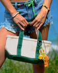 A person in frayed denim shorts holds a Venessa Arizaga canvas tote with green straps, colorful beaded bracelets, and a playful Bag Key Leash keychain. Green grass and blue sky complete the scene.