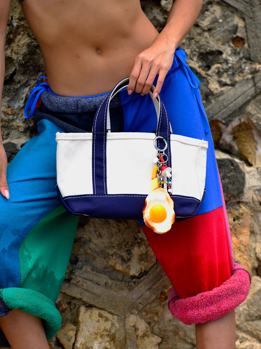 A person in colorful patchwork pants and no shirt holds a Venessa Arizaga Bag Key Leash attached to a white and navy tote bag, standing before a stone wall.