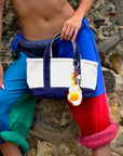 A person in colorful patchwork pants and no shirt holds a Venessa Arizaga Bag Key Leash attached to a white and navy tote bag, standing before a stone wall.