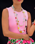 A woman in a sleeveless pink top and floral skirt wears the Venessa Arizaga Vibe Check Necklace 30-38", layered with pearls and colorful charms, plus a red bracelet, posing against a black background.