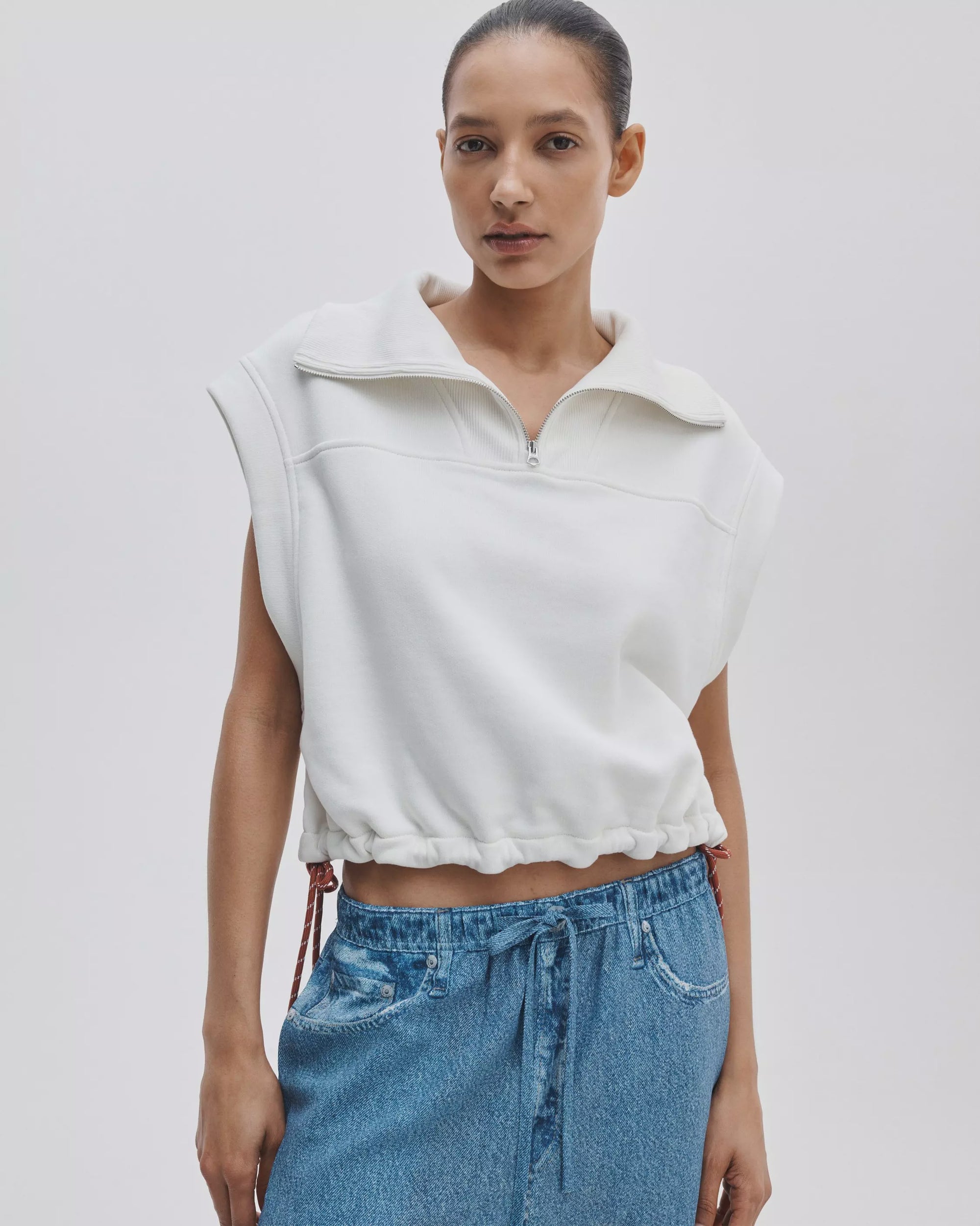 A woman stands against a plain background, confidently wearing the Rag & Bone vintage terry half zip muscle tee with a collar and drawstring hem, paired with blue drawstring denim jeans. Her hair is pulled back as she looks at the camera.