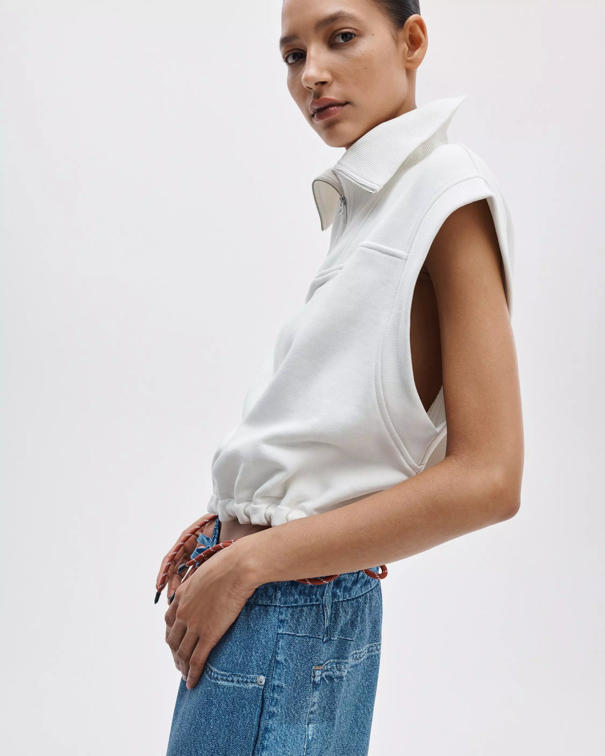 A person in a vintage look stands against a plain white background, wearing high-waisted blue jeans and the Rag & Bone vintage terry half zip muscle tee, gazing at the camera with a relaxed pose.