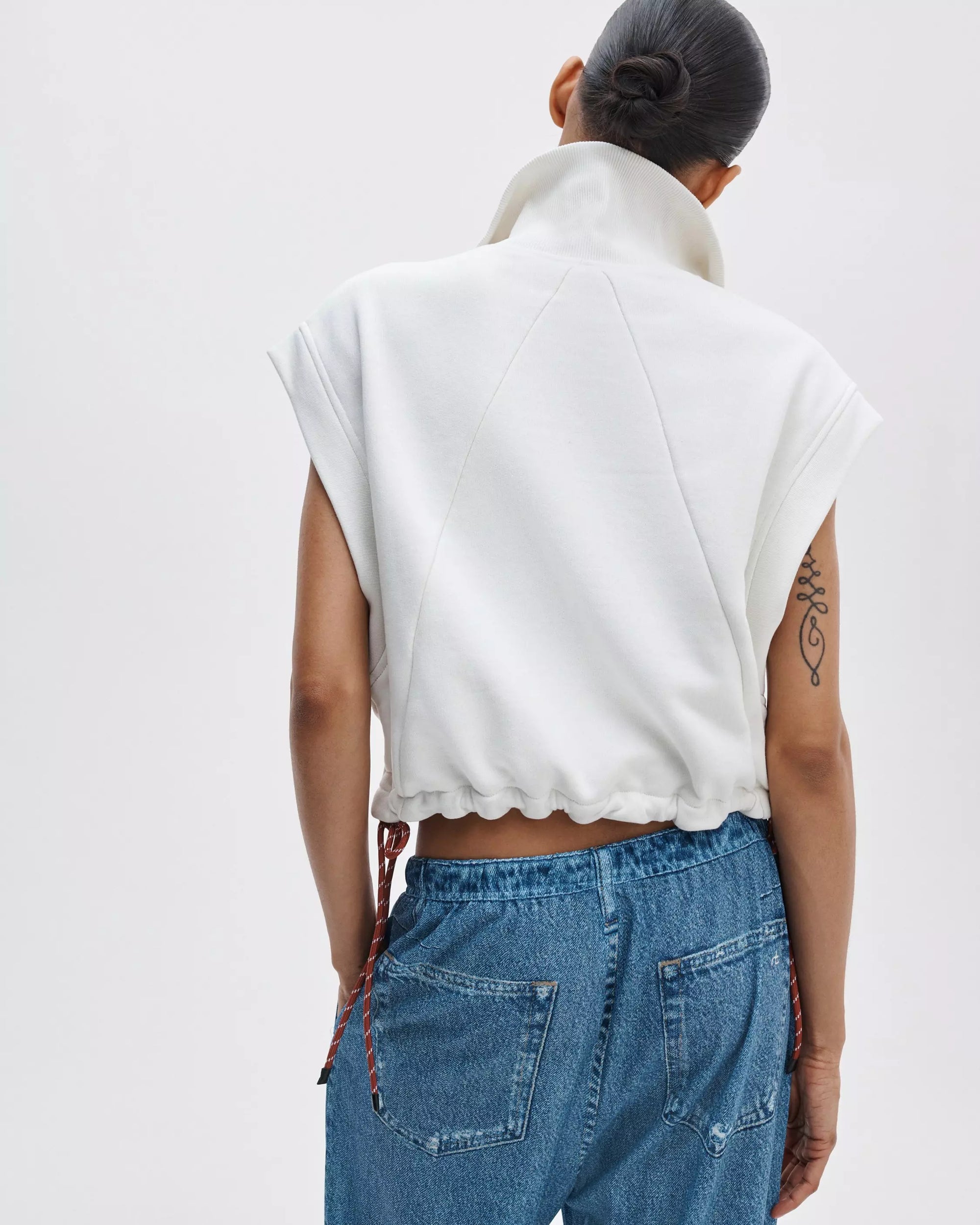 A person with their hair in a bun is seen from behind, wearing Rag & Bone's vintage terry half zip muscle top in white and blue jeans. A tattoo decorates their upper left arm.