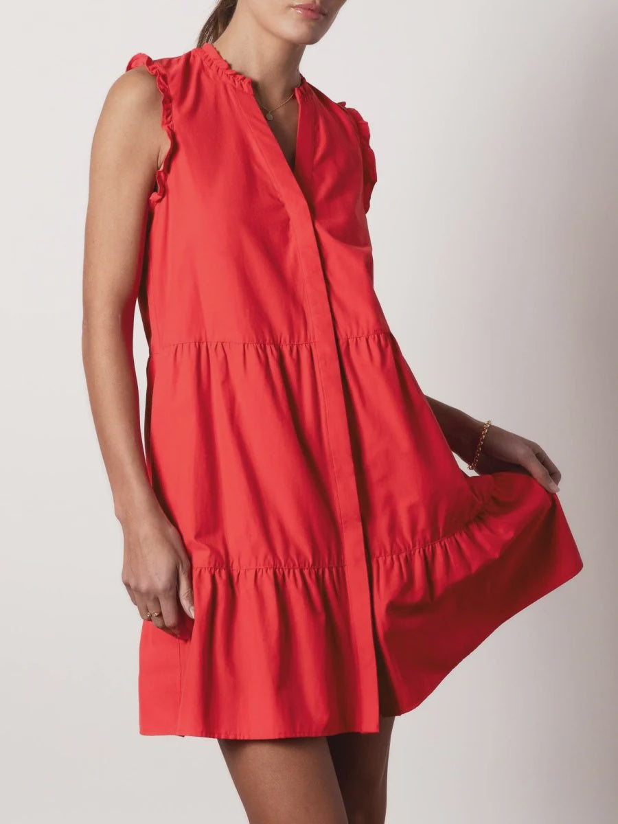 A woman wears the sleeveless VIOLA DRESS by BROCHU WALKER, featuring a bright red hue, V-neckline, tiered mini skirt, and holds the skirt out to the side against a plain light background.