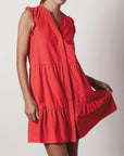 A woman wears the sleeveless VIOLA DRESS by BROCHU WALKER, featuring a bright red hue, V-neckline, tiered mini skirt, and holds the skirt out to the side against a plain light background.