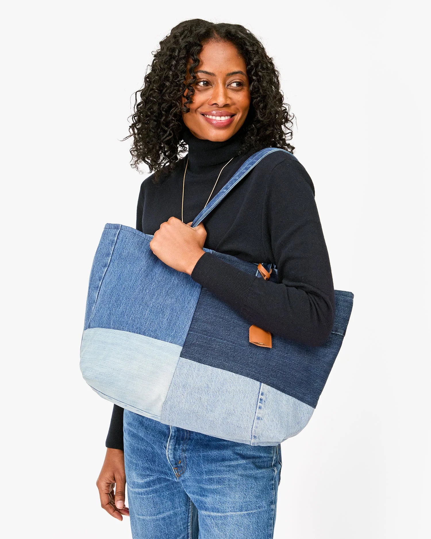 A person with curly hair wears a black turtleneck and blue jeans, holding the Volie bag by Clare Vivier, crafted from up-cycled jeans. They smile against a plain white background.