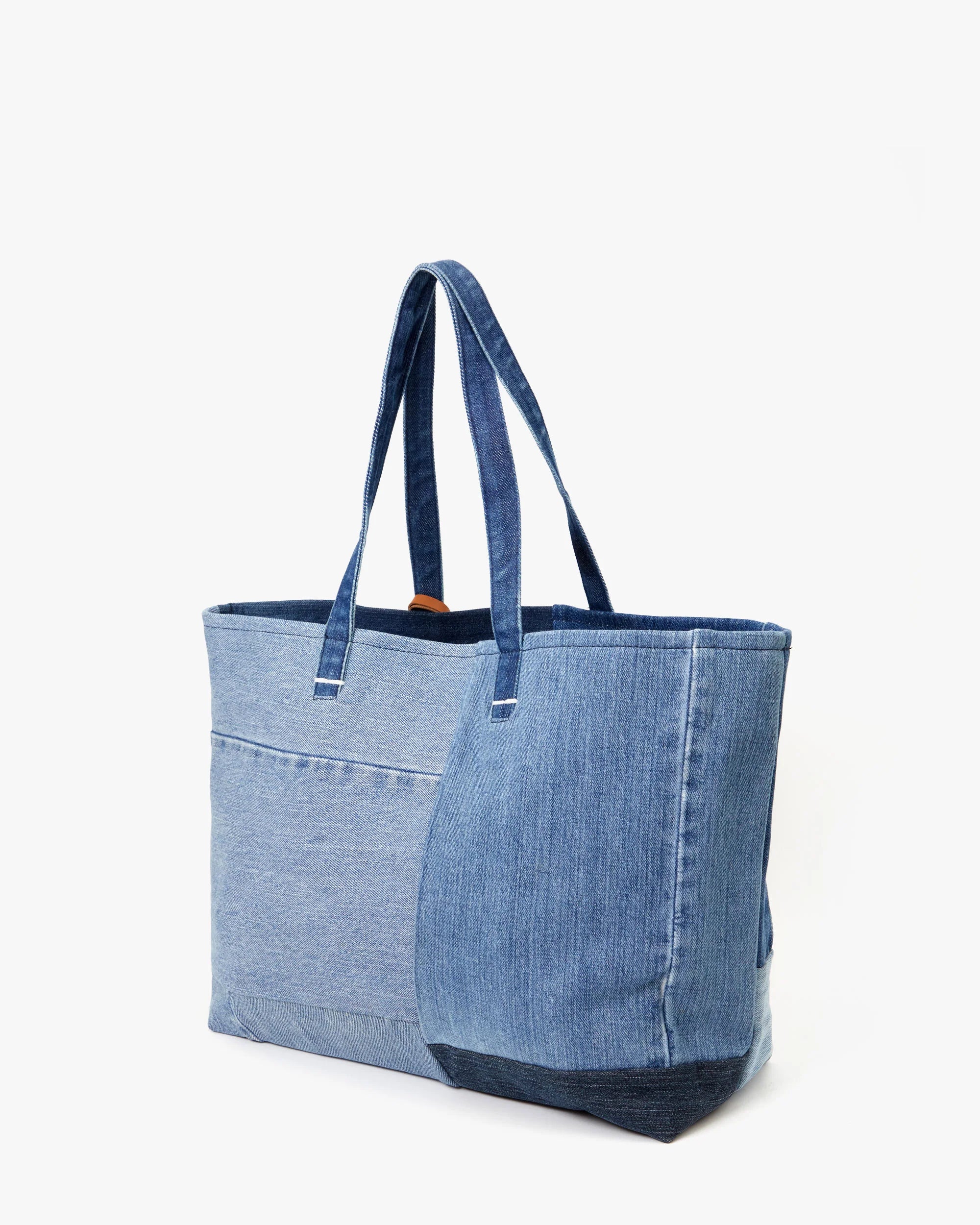 The Volie by Clare Vivier is an up-cycled jeans tote bag with dual blue fabrics and two shoulder straps. It has a unique front pocket and stands upright against a white background.