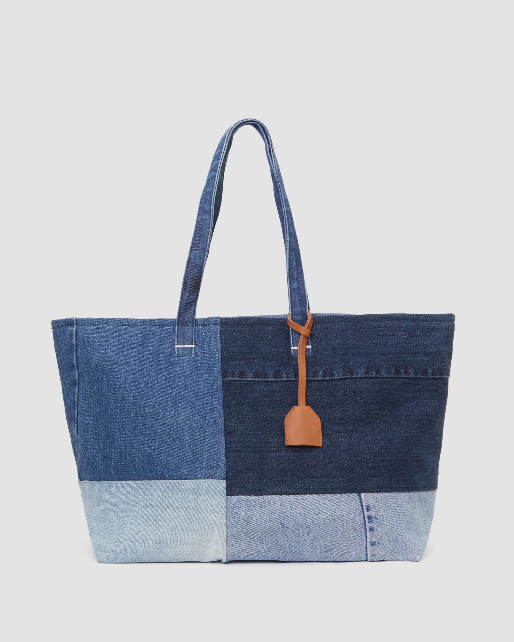 Clare Vivier's Volie is a large tote bag made from up-cycled jeans, showcasing a patchwork design with varying blue patches. It has two denim handles and a brown vegetable-tanned leather tag on one strap, displayed against a plain white background. Made in L.A.