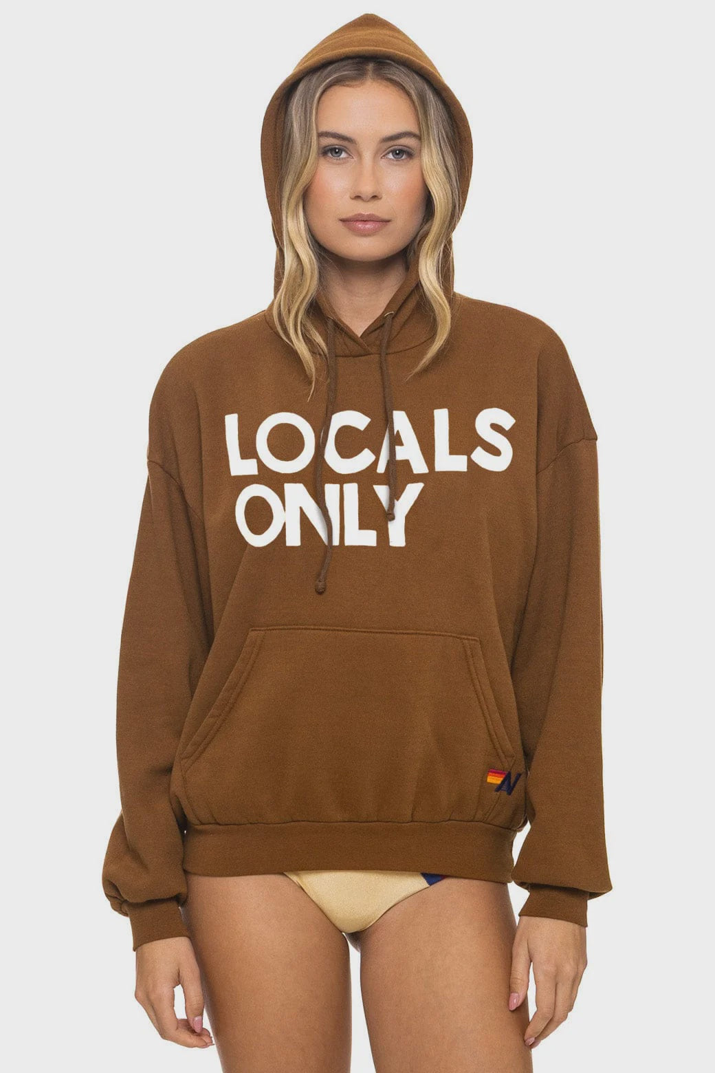 A model stands against a plain white background in Aviator Nation’s “locals only - pullover hoodie relaxed” with “LOCALS ONLY” in bold white letters, hood up, paired with beige bikini bottoms.