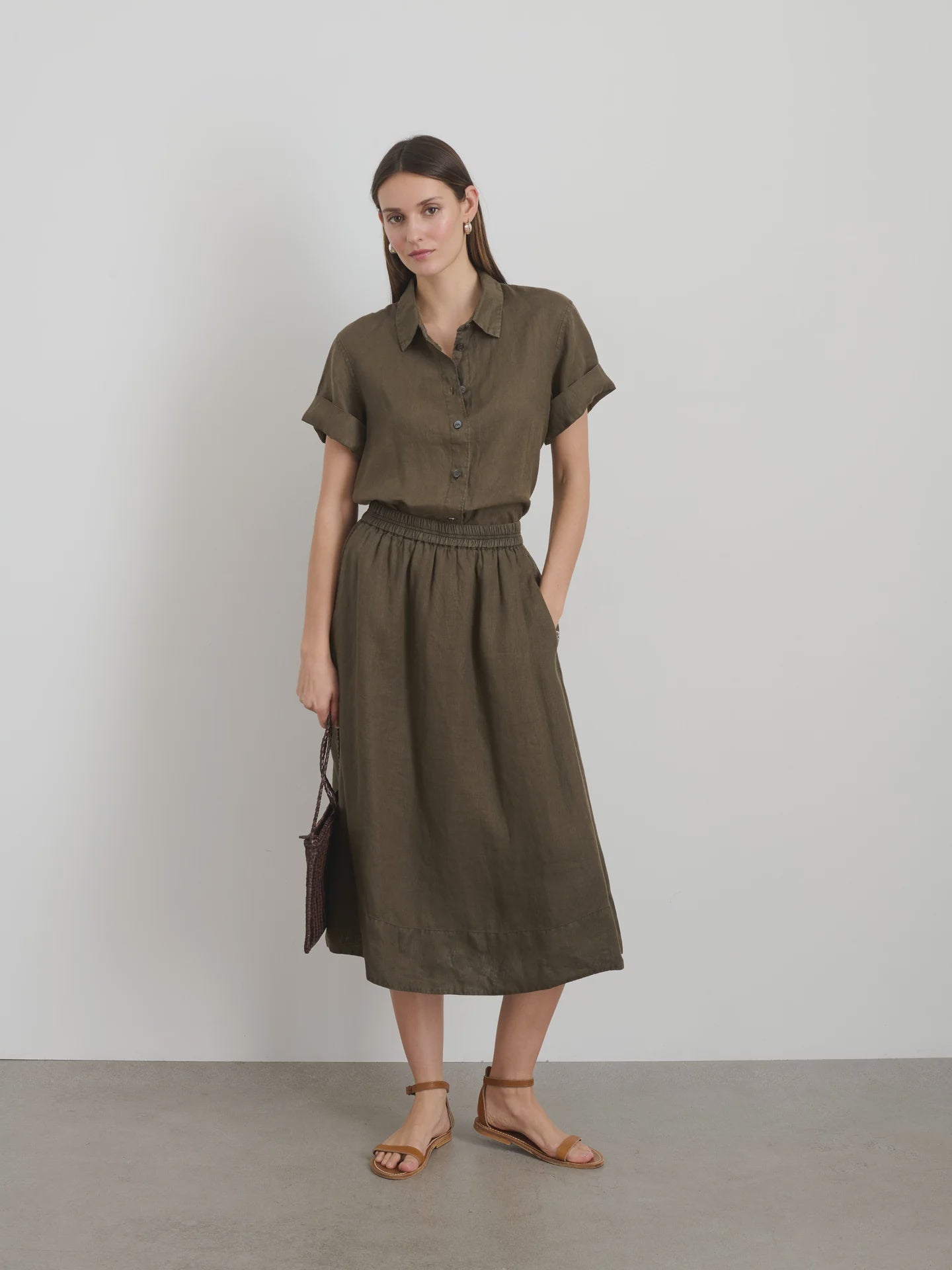 Against a plain white wall, a person wears an olive-green garment-dyed button-up shirt and the Maddie Skirt Linen by Alex Mill. They hold a small brown bag and wear brown sandals, gazing softly at the camera. The scene is minimalistic.