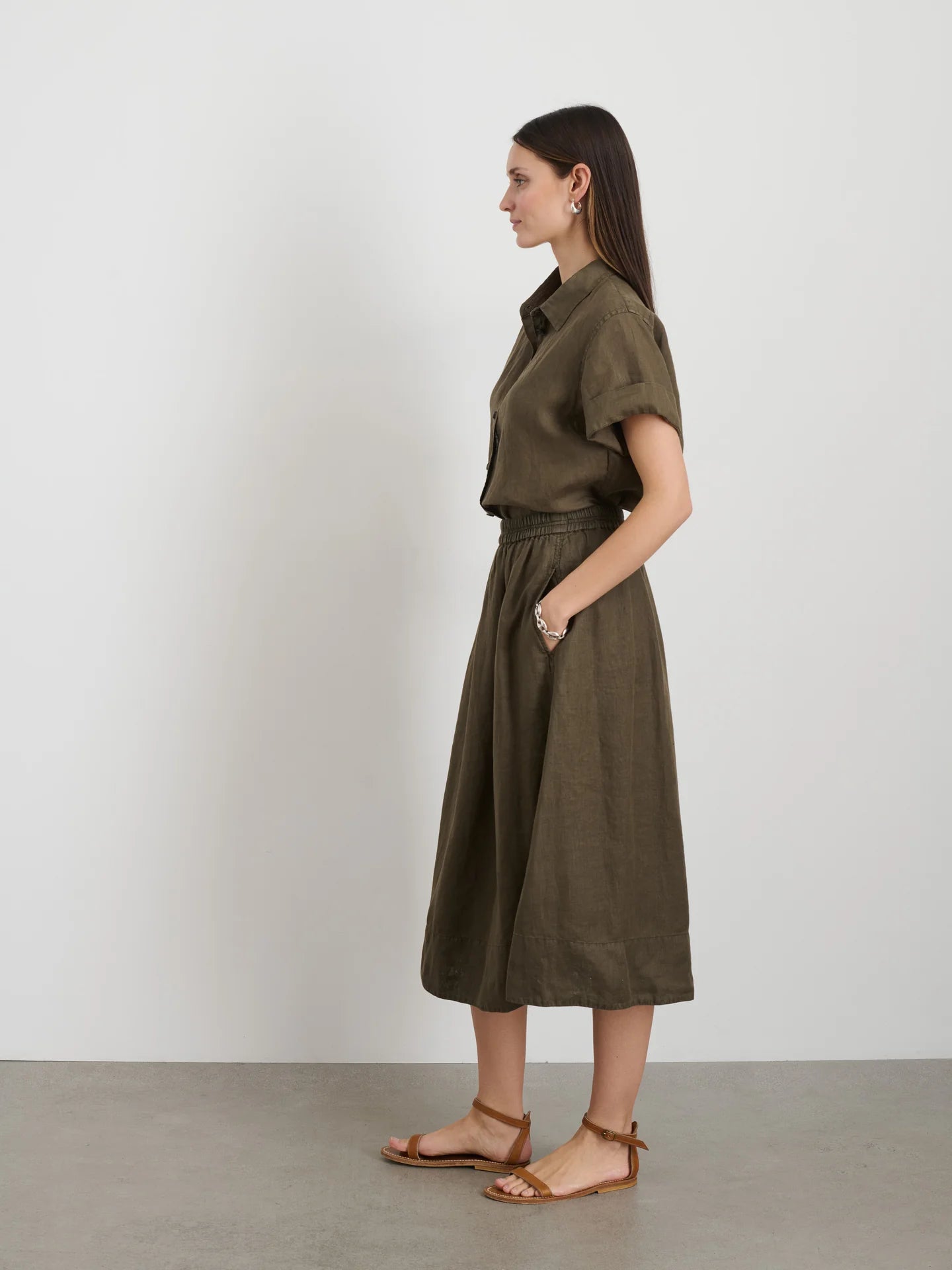 A side profile showcases the garment-dyed olive green "Maddie Skirt Linen" by Alex Mill featuring iPhone-sized pockets and a gathered waist. Long, straight hair flows as they wear brown sandals against a plain white wall on a gray floor.