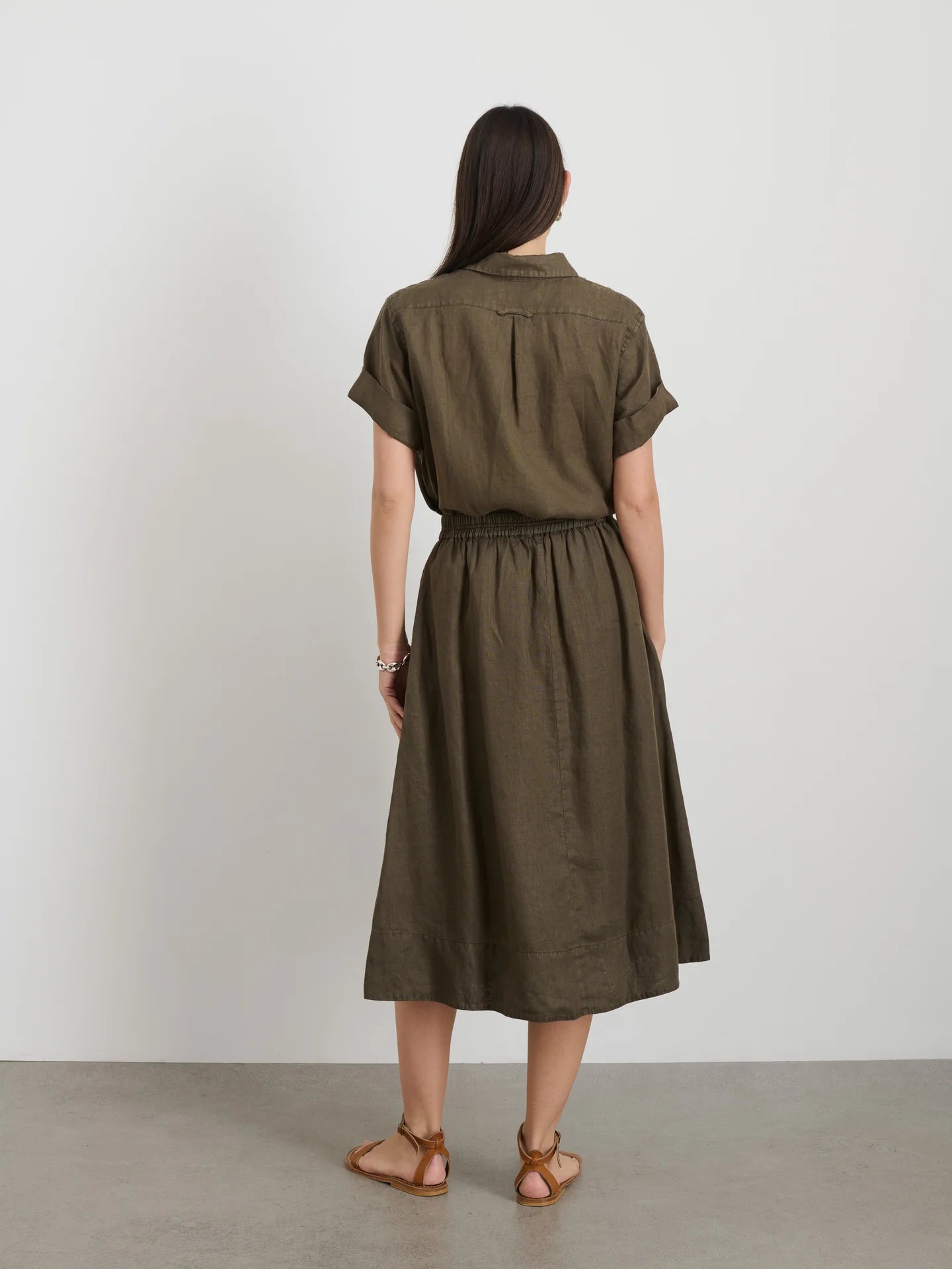 A person with long dark hair is barefoot on a light floor, facing a plain white wall, wearing the Alex Mill Maddie Skirt Linen in olive green. The skirt is knee-length with short sleeves and iPhone-sized pockets.