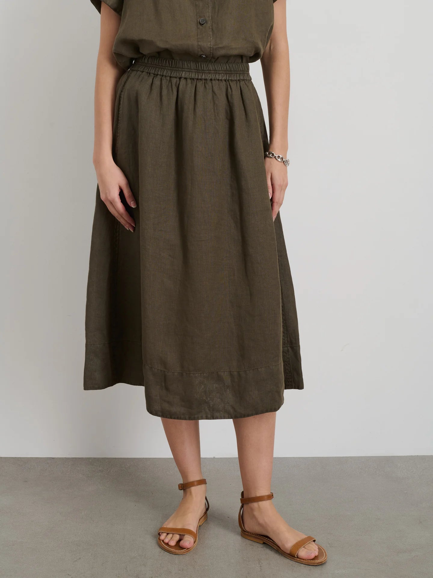 Outfitted in Alex Mill's Maddie Skirt Linen, a knee-length olive green garment with iPhone-sized pockets, paired with a matching sleeveless top. The look is completed with brown sandals and a bracelet against a plain light gray wall.